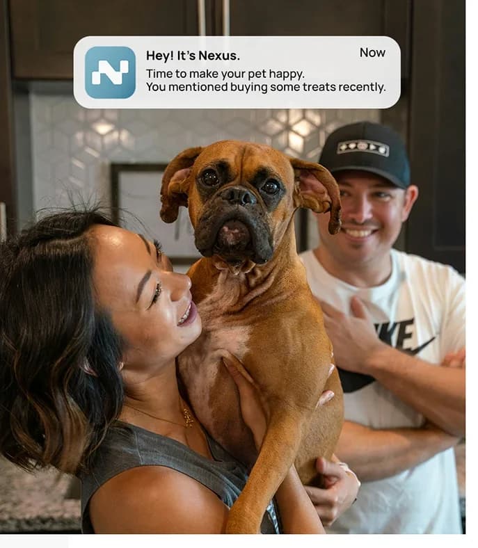 A couple in a kitchen holding up their boxer dog, laughing, under a Nexus notification reading “Time to make your pet happy — you mentioned buying some treats recently”
