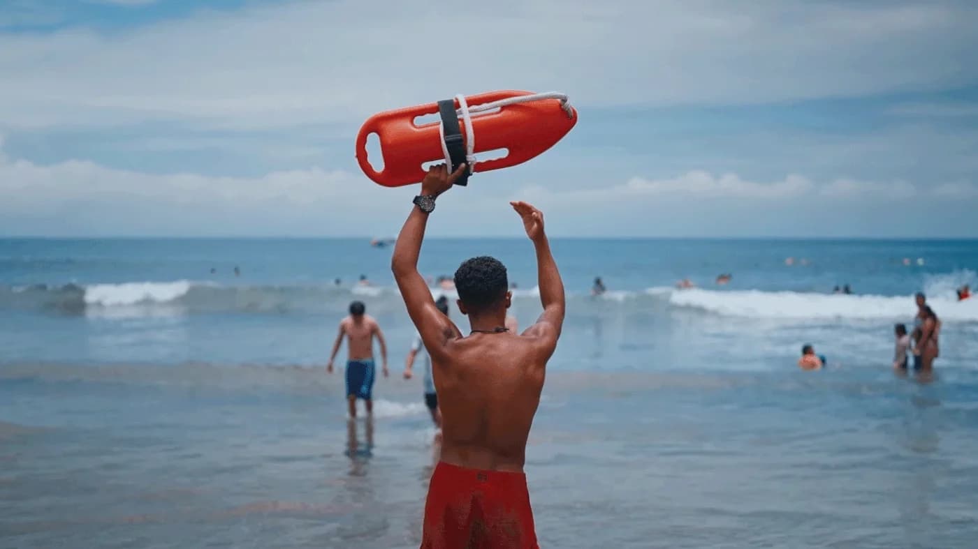 A lifeguard holding a rescue can overhead at the water’s edge, swimmers in the surf beyond