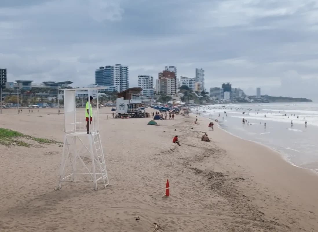 An unbranded white lifeguard tower standing on a wide stretch of beach at Manta, the city skyline behind it and bathers in the surf