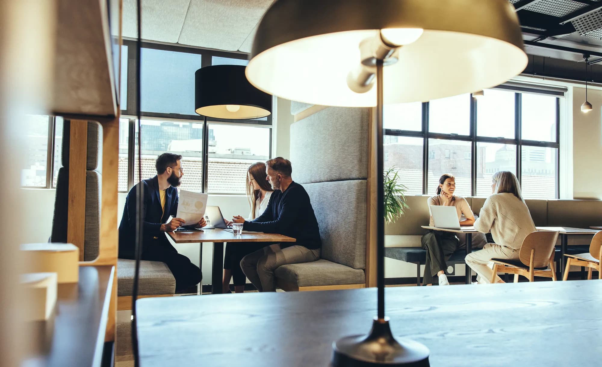 An open-plan office with two groups working at laptops in booth seating by tall windows