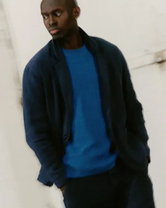 A man in a navy linen overshirt worn open over a cobalt waffle knit, against a plaster wall