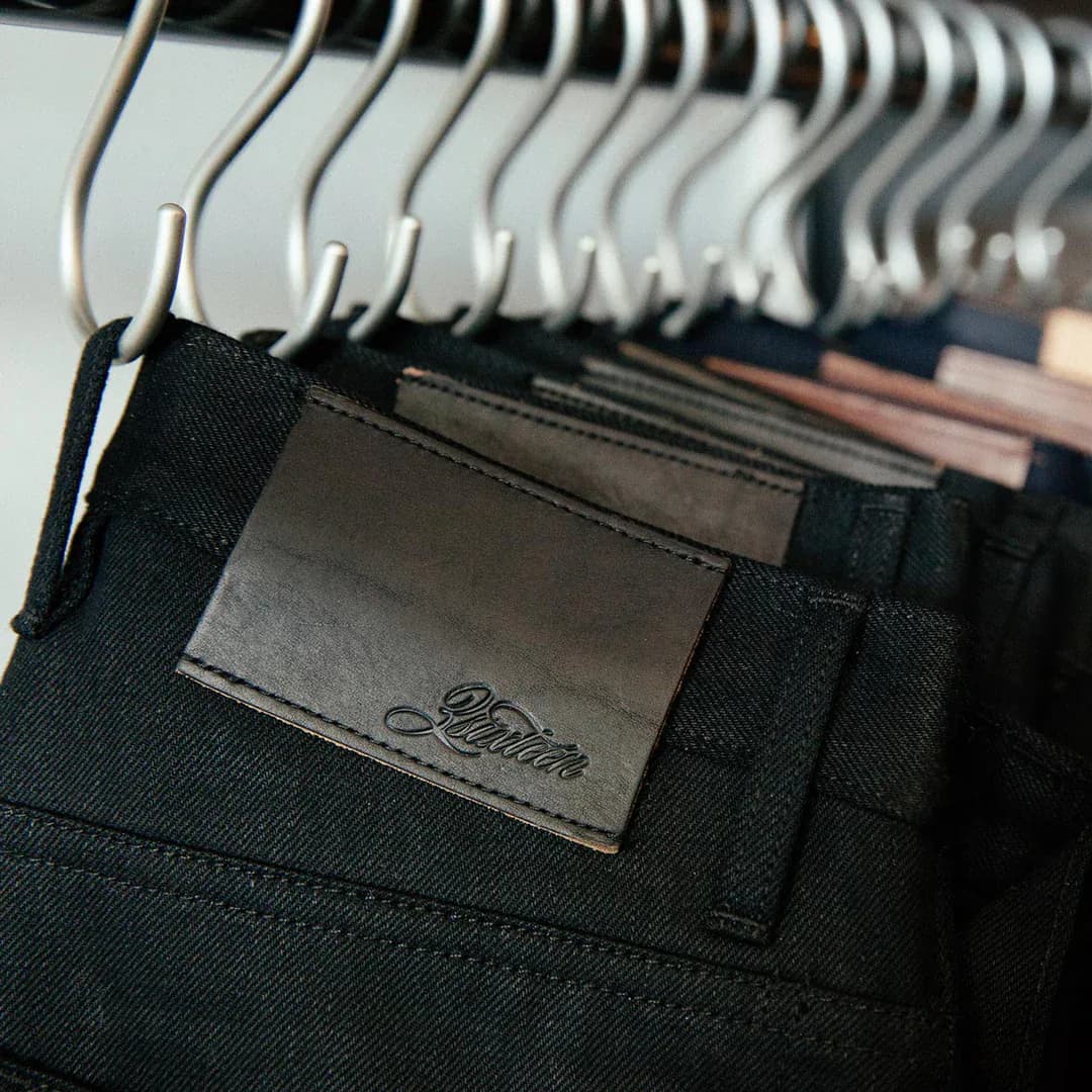 A rack of black denim on steel hangers, the front pair showing a blind-embossed 3sixteen leather patch