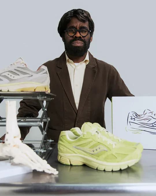 A designer in a brown jacket beside the 3sixteen and Saucony Guide 7 collaboration, a sketch and an anatomical foot model on the table