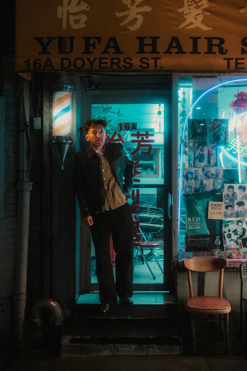A man in a leather-and-denim jacket standing in the neon-lit doorway of a barbershop on Doyers Street, Chinatown