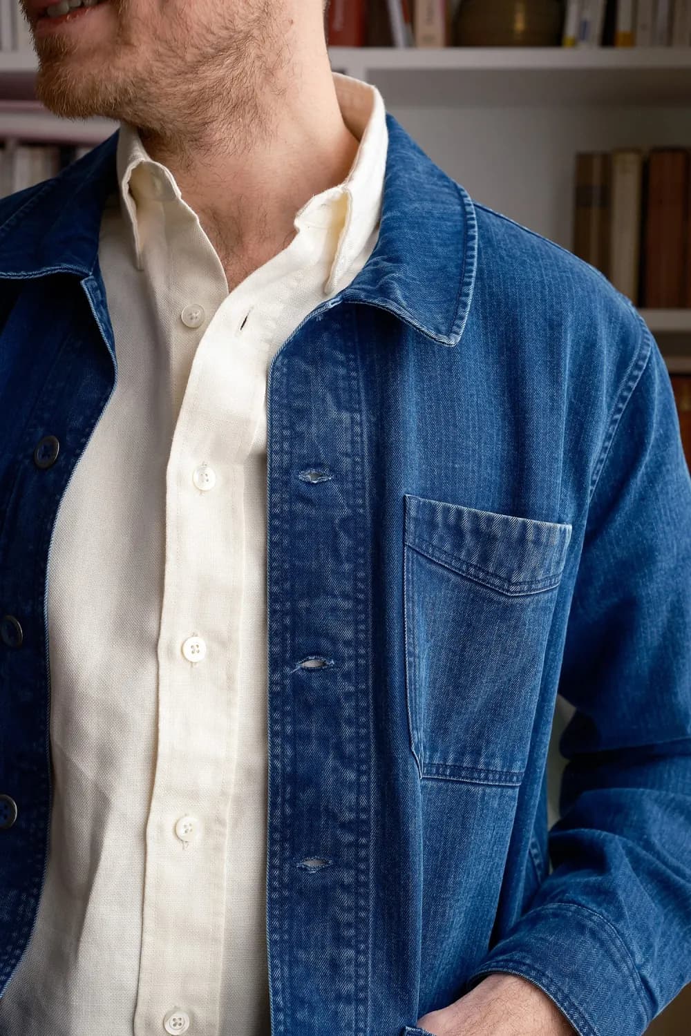 Close detail of a faded indigo chore coat worn open over a cream oxford shirt, bookshelves behind