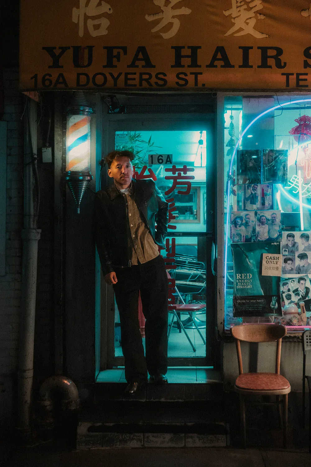 A man in a leather-and-denim jacket standing in the neon-lit doorway of a barbershop on Doyers Street, Chinatown