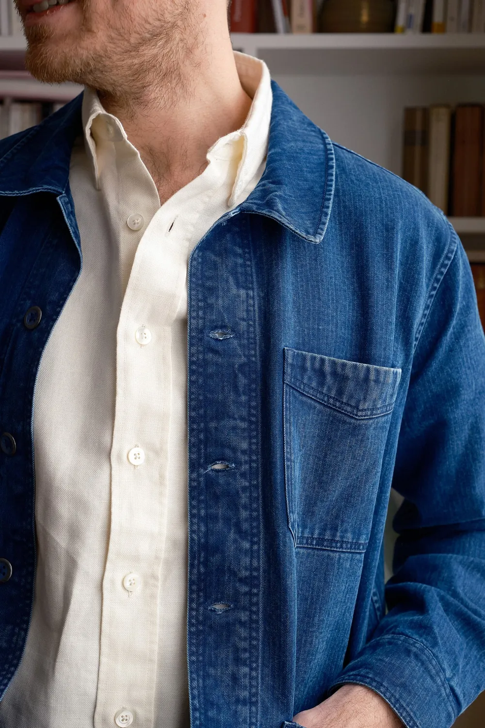 Close detail of a faded indigo chore coat worn open over a cream oxford shirt, bookshelves behind