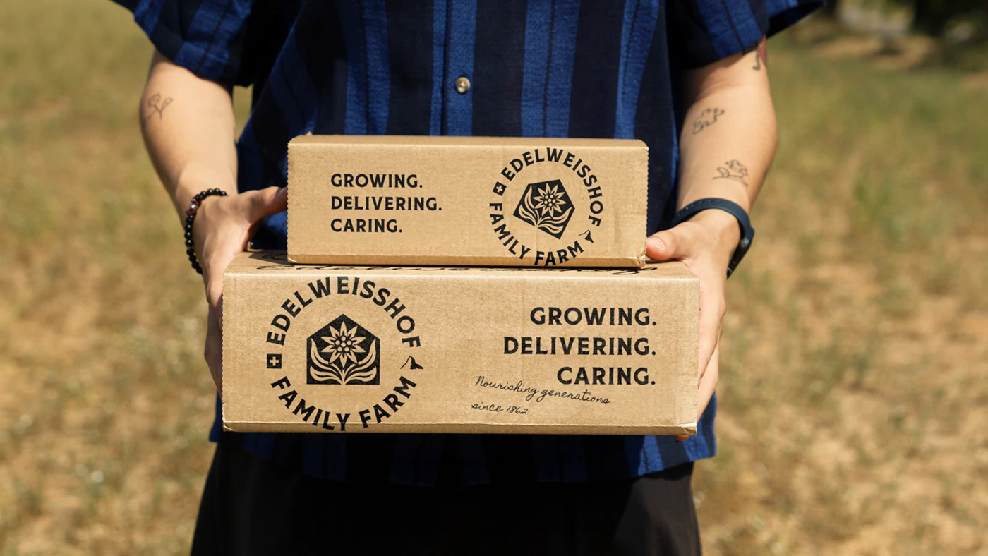 Two kraft Edelweisshof shipping boxes carried against a sunlit field, printed in single-colour black with the crest and the line “Growing. Delivering. Caring.”