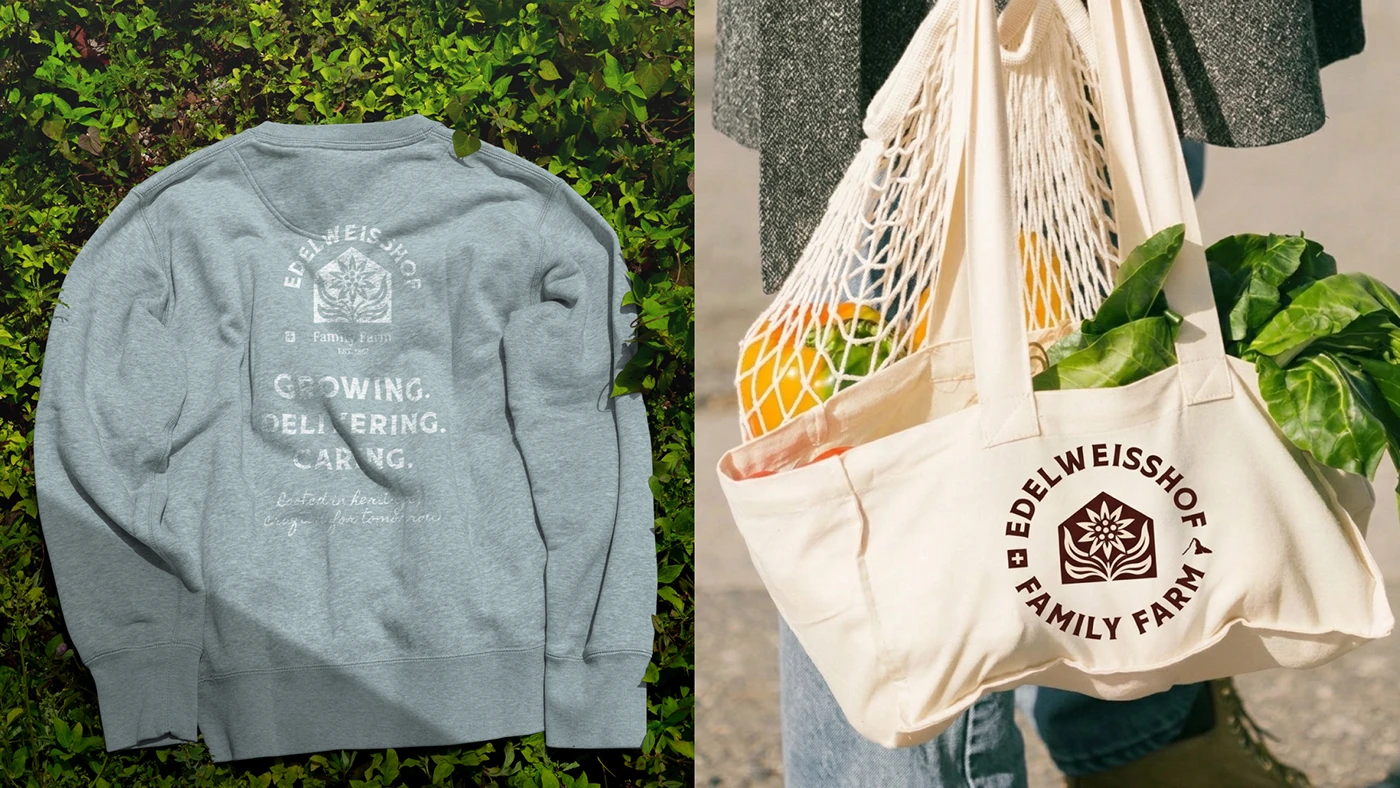 A grey Edelweisshof sweatshirt printed “Growing. Delivering. Caring.” lying on foliage, beside a canvas market tote of oranges and chard carried on a cobbled street