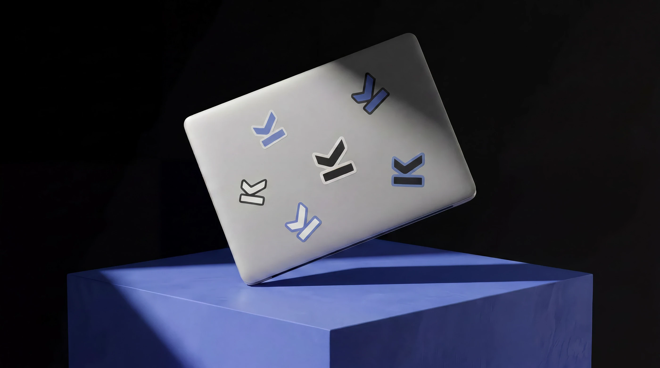 Laptop stickers of the Mark OS logomark in blue, black and white, arranged on a silver lid above a blue plinth