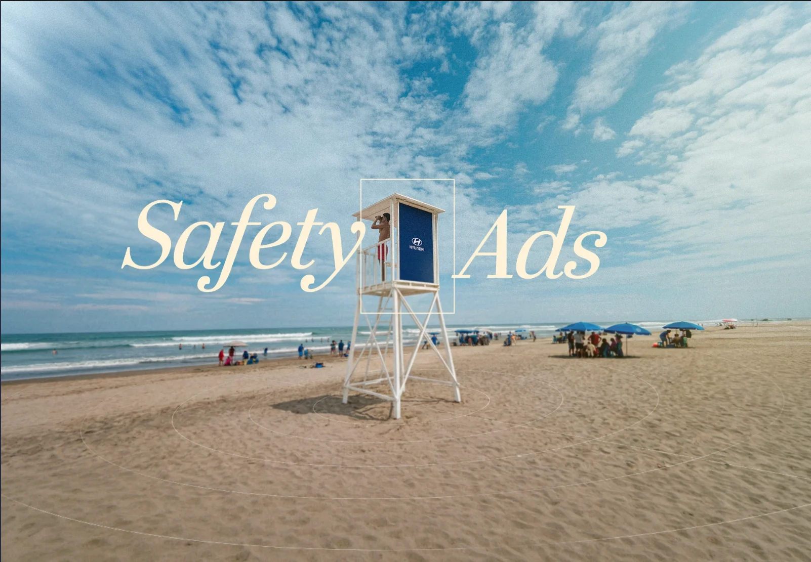 Safety Ads: a lifeguard tower carrying a Hyundai panel stands on a wide Manta beach under a high sky, a lifeguard scanning the water from its deck