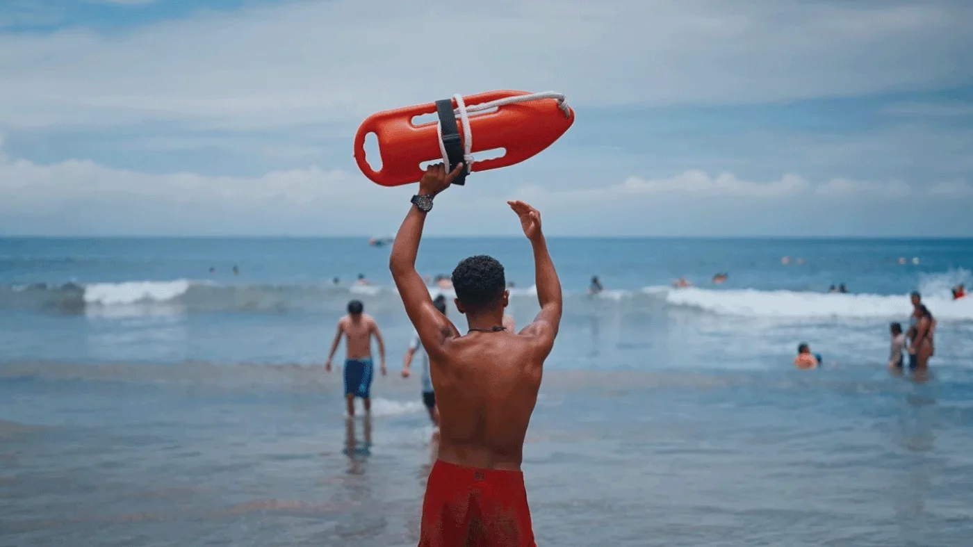 A lifeguard holding a rescue can overhead at the water’s edge, swimmers in the surf beyond