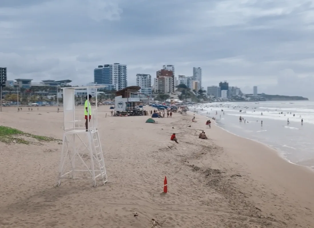 An unbranded white lifeguard tower standing on a wide stretch of beach at Manta, the city skyline behind it and bathers in the surf