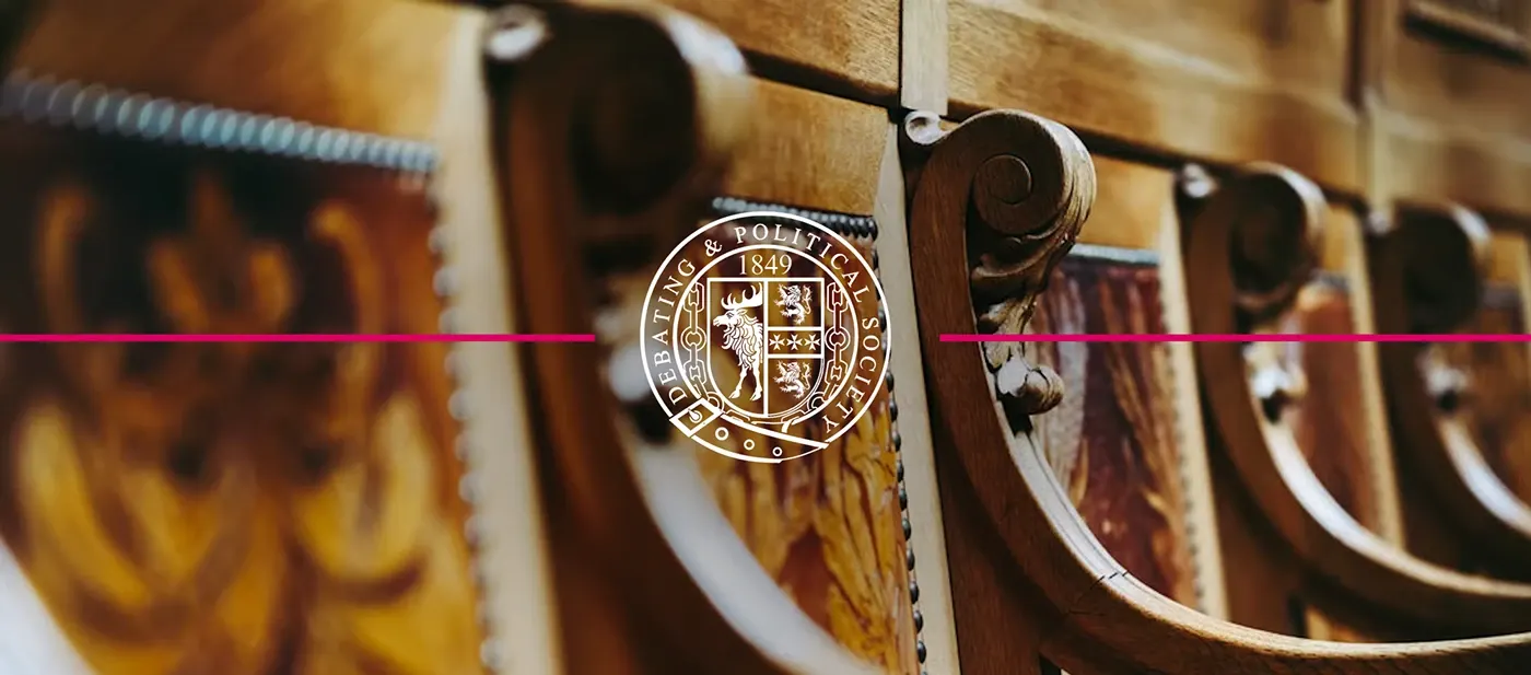 Carved wooden chamber seats in warm light, overlaid with a circular debating society crest.