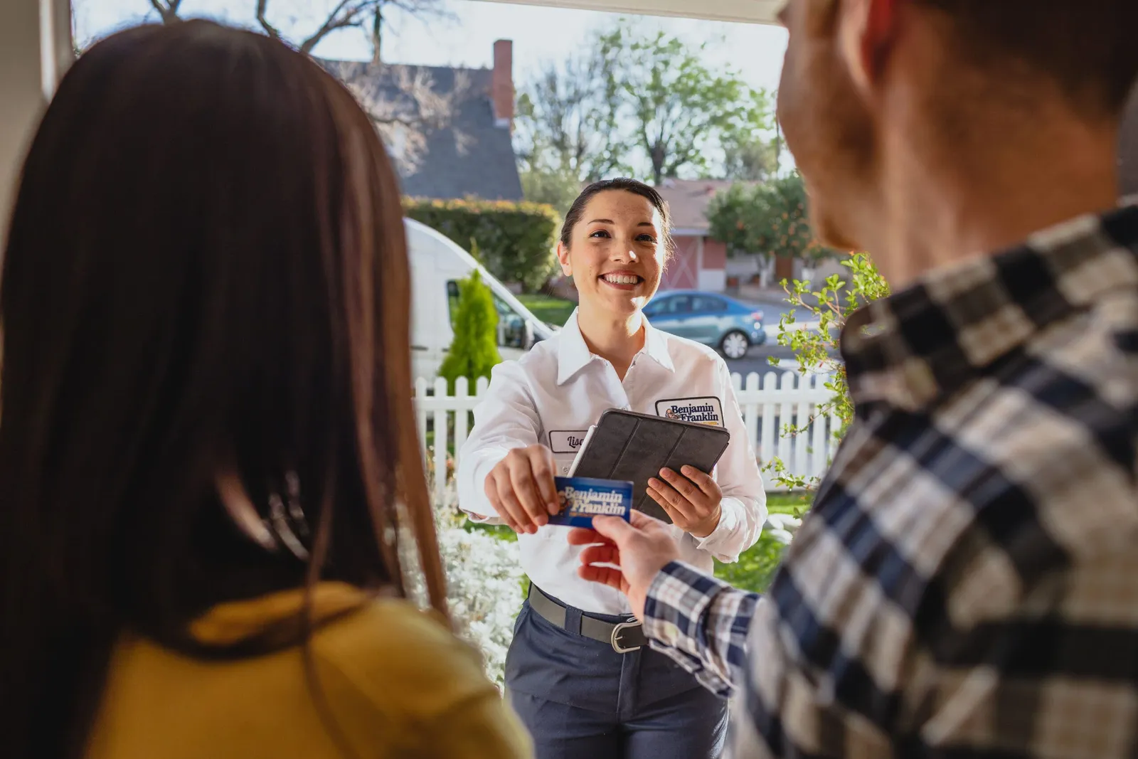 Benjamin Franklin Plumbing technician in uniform at a front door, handing a branded card to two homeowners