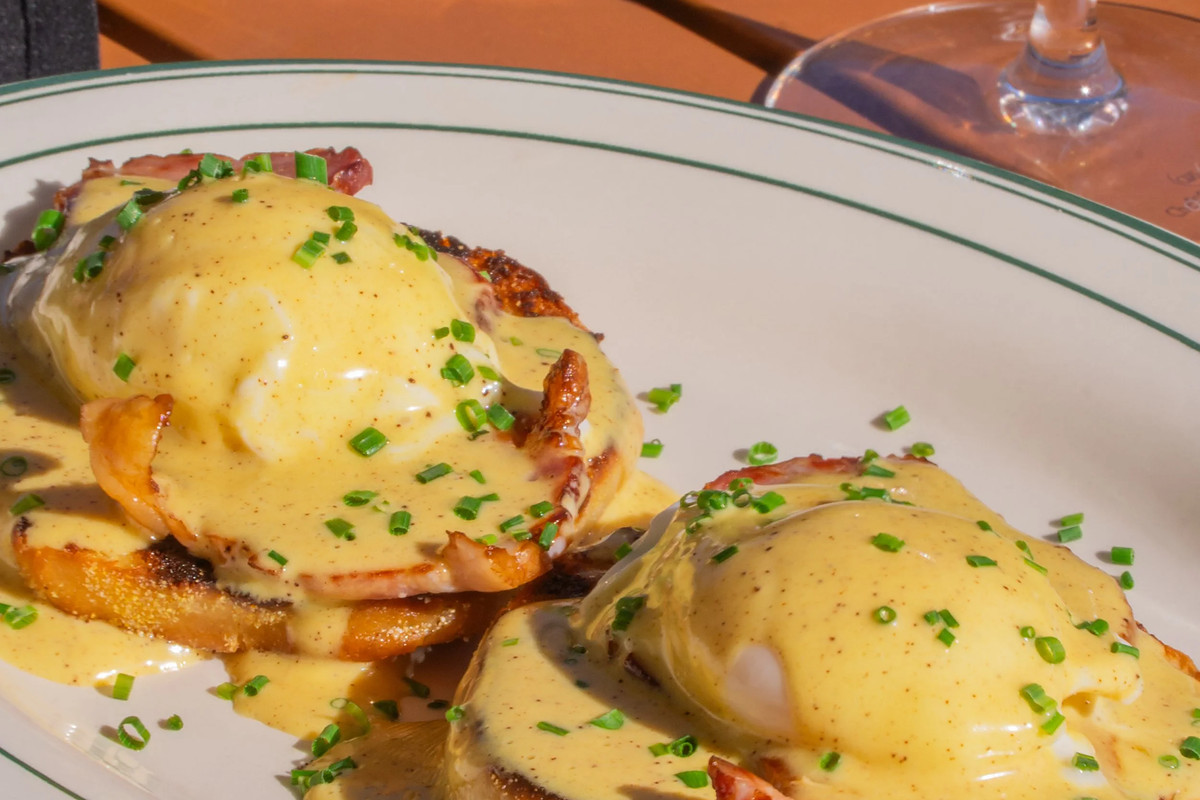 Two eggs Benedict on toasted English muffins with ham and hollandaise, scattered with chives, on a green-rimmed plate in the sun.