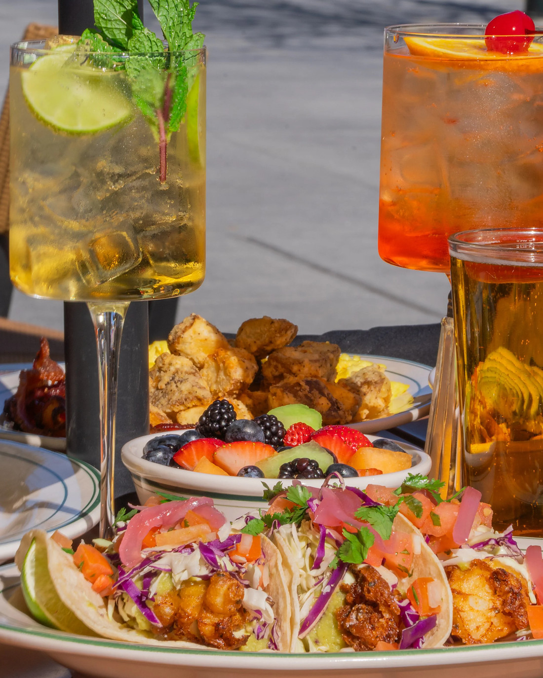 Two cocktails garnished with mint, lime and a cherry beside a glass of beer, with fish tacos and fried potatoes on the table behind them.