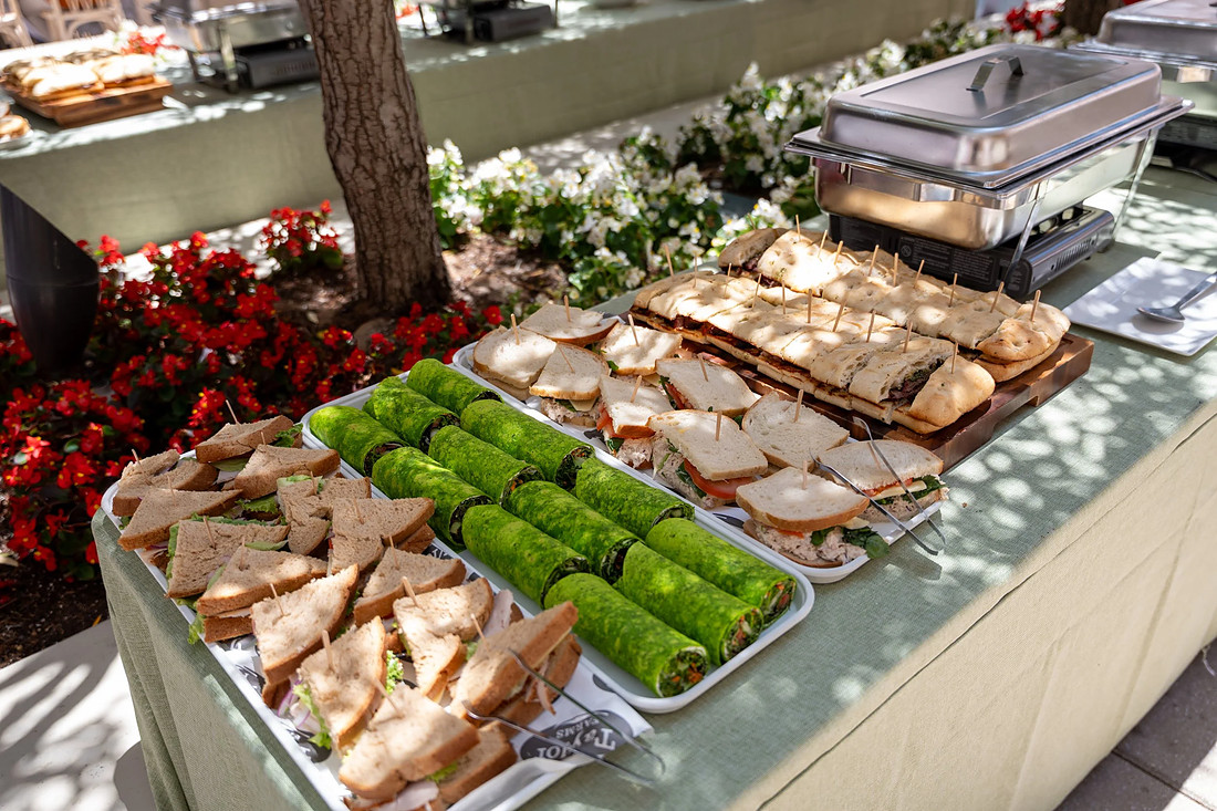 An outdoor buffet line: trays of quartered sandwiches and green wraps beside chafing dishes, set out on a table by a flowering bed.