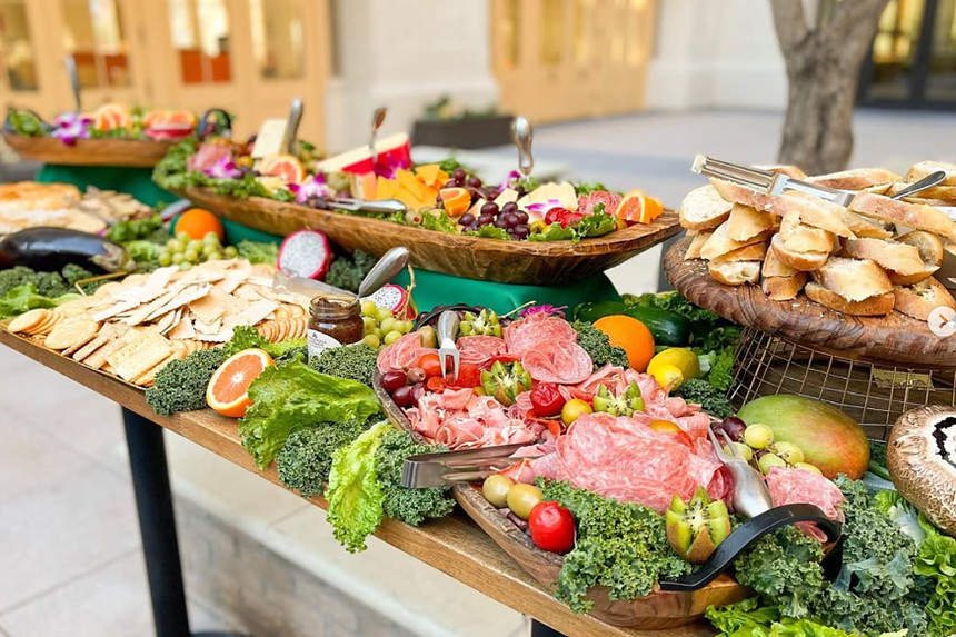 A grazing table set outdoors: wooden boards of cured meats, cheeses, olives, sliced fruit and baguette, banked with kale and edible flowers.