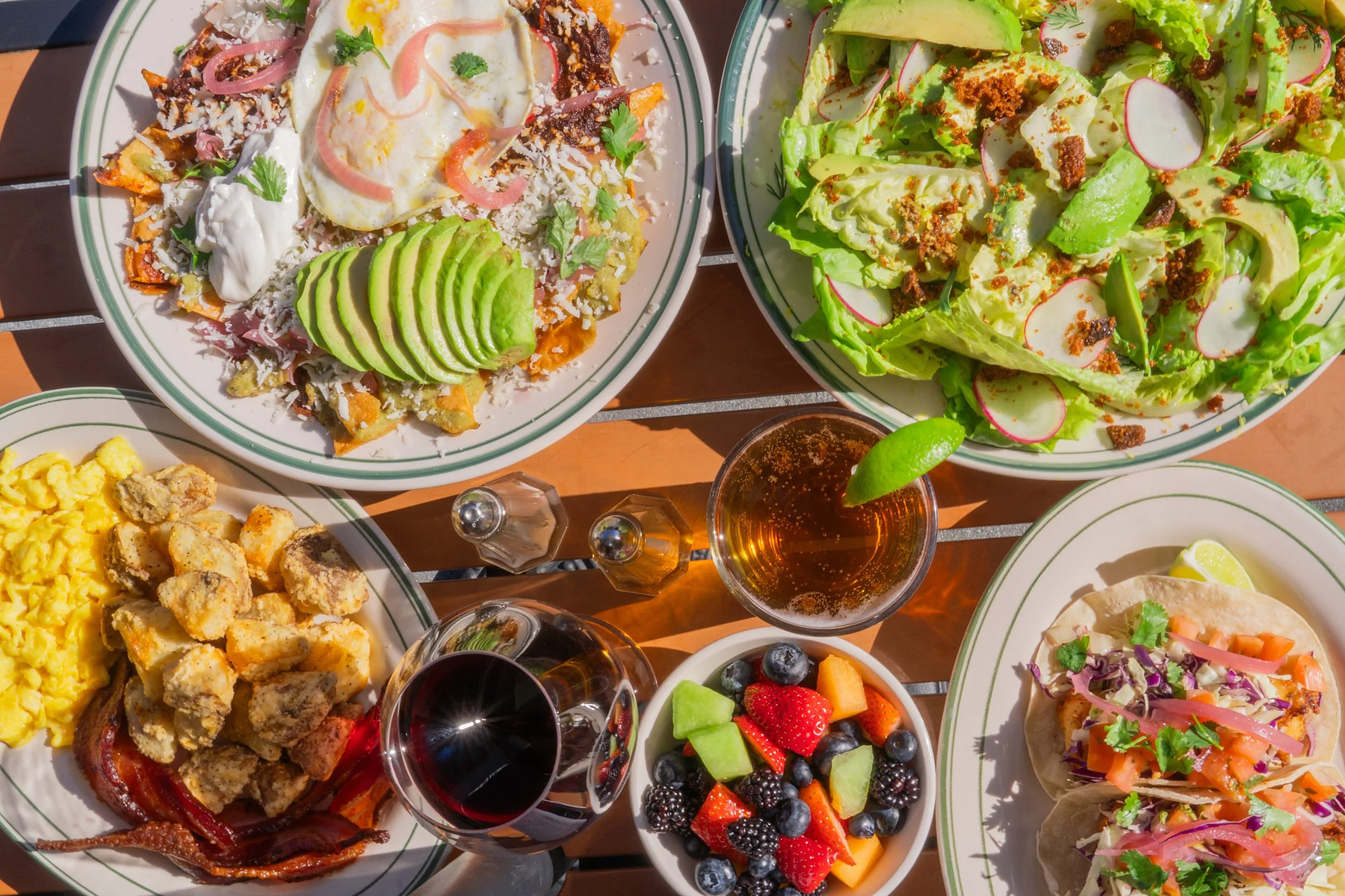 An overhead table on the patio: chilaquiles under a fried egg with sliced avocado, a gem lettuce salad with radish, scrambled eggs with bacon and potatoes, fish tacos, a bowl of berries and melon, a glass of red wine and a beer.