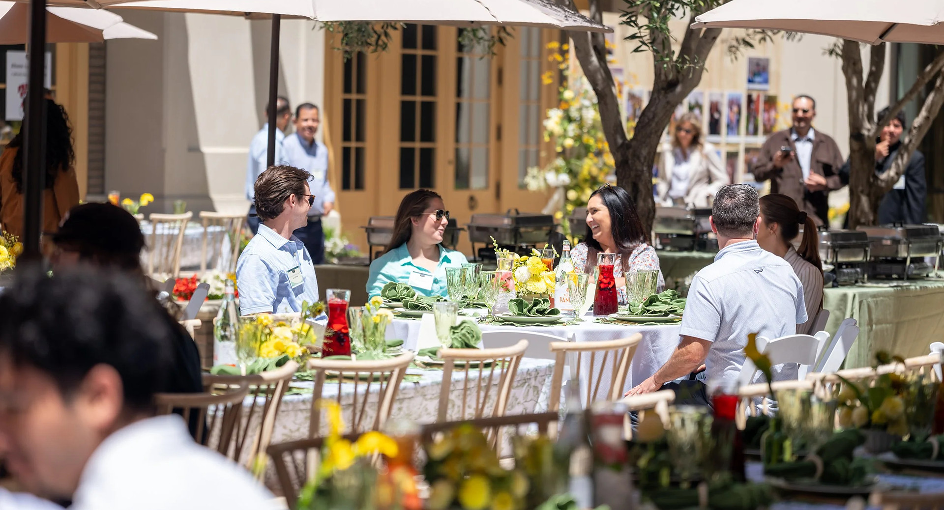 Guests seated at round tables on the Portobello's patio under cream umbrellas, with olive trees, yellow flowers and folded green napkins on the linen.