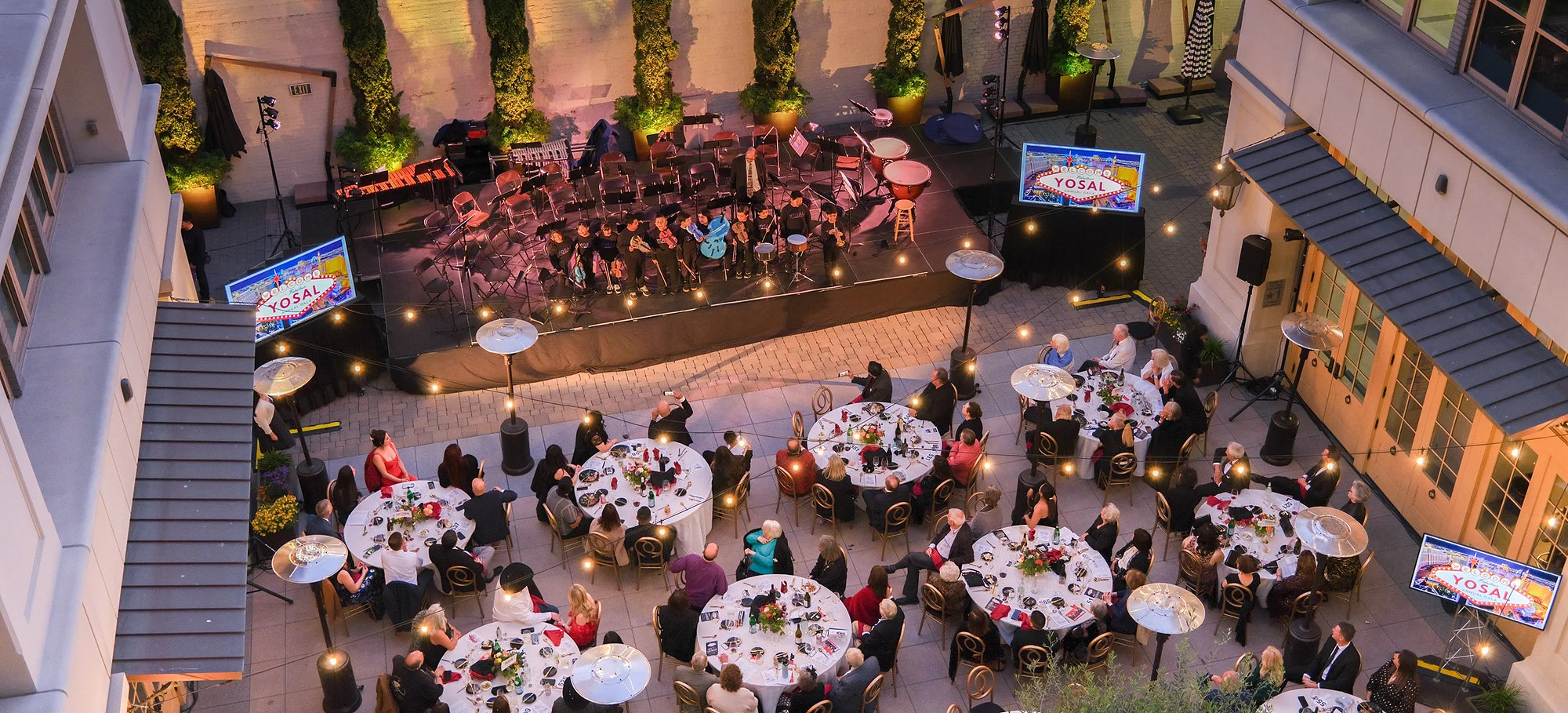 The patio from above after dark, round tables laid and lit under the trees, guests seated across the courtyard.