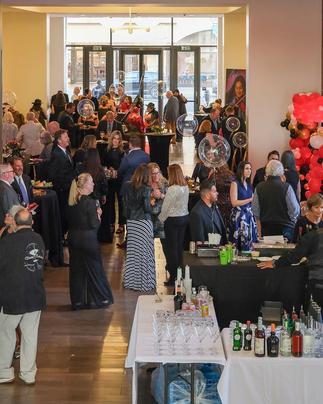 Inside the restaurant during an event: guests standing along the bar with bottles and glassware set out, tall windows onto Main Street behind them and a red floral arrangement by the door.