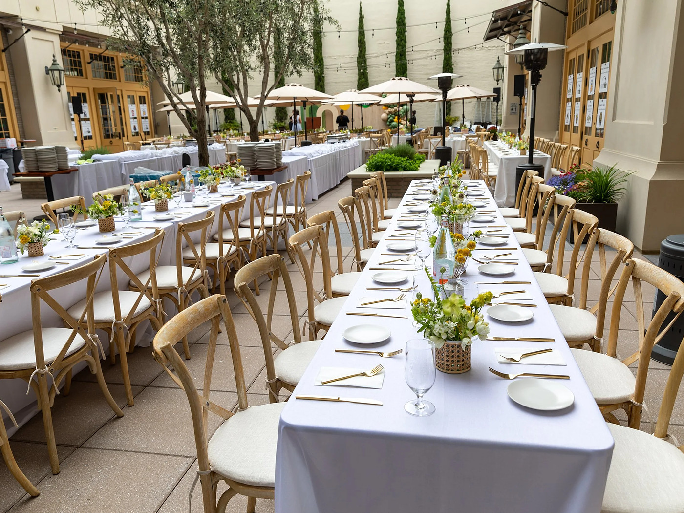 Long banquet tables laid end to end on the patio under white linen, with cross-back chairs down both sides, small green centrepieces and cypress trees behind.